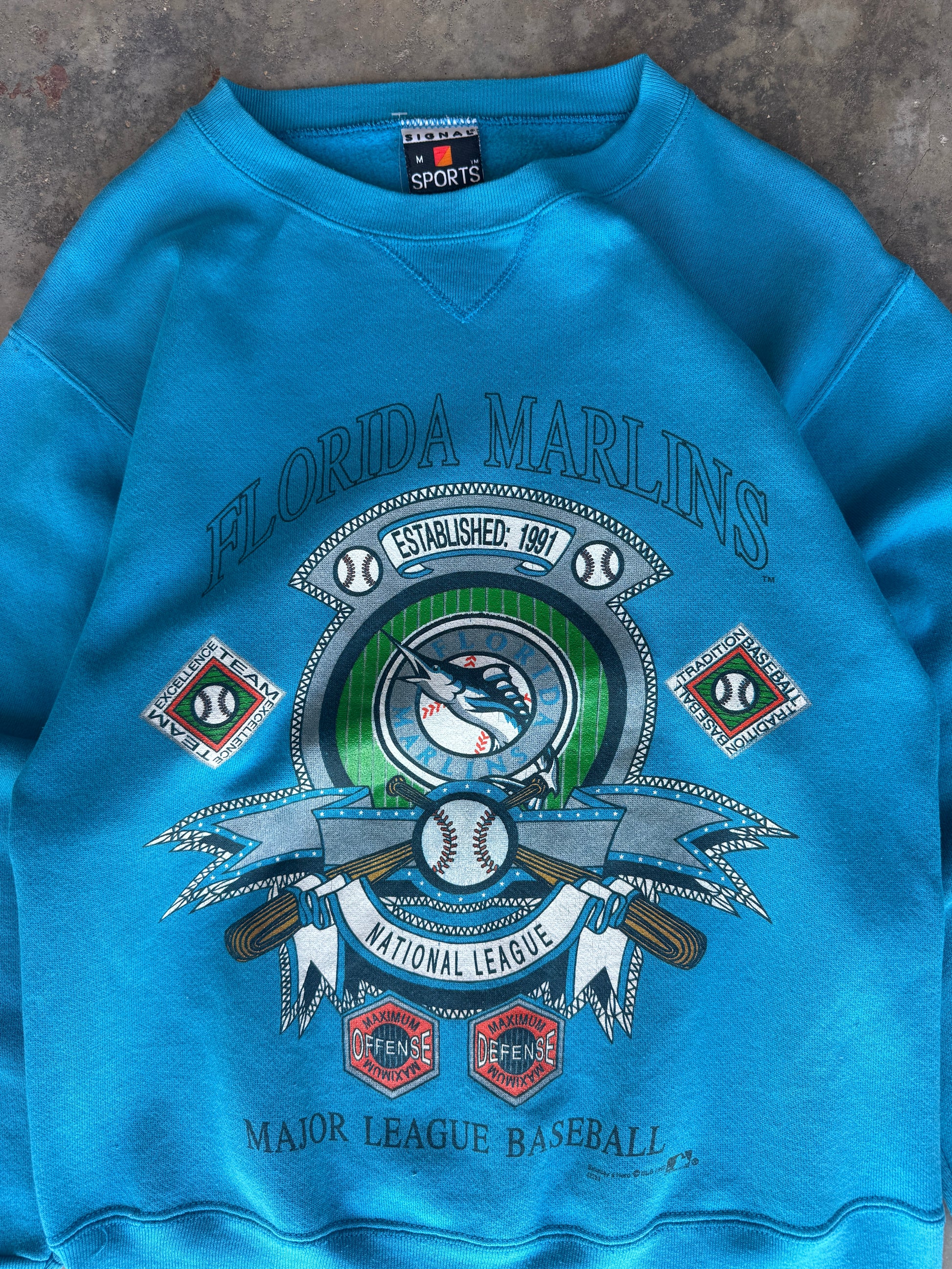 A bright blue crewneck sweatshirt featuring a large Florida Marlins National League graphic on the front, laid flat on a textured gray surface.