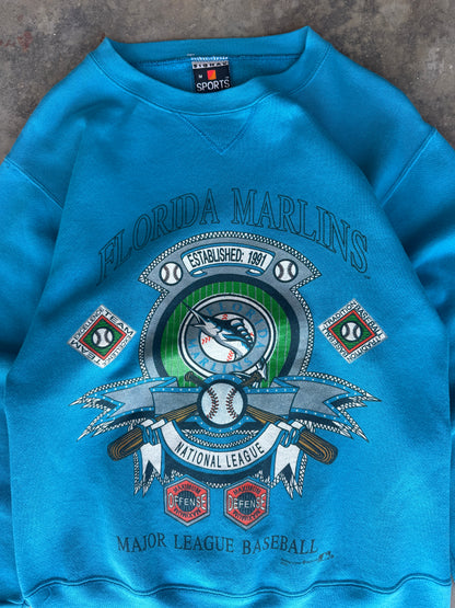 A bright blue crewneck sweatshirt featuring a large Florida Marlins National League graphic on the front, laid flat on a textured gray surface.