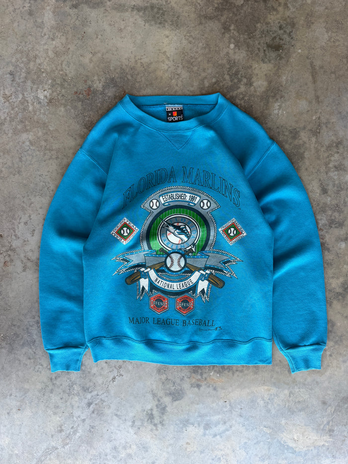 A bright blue crewneck sweatshirt featuring a Florida Marlins graphic print, laid flat on a textured gray concrete surface.
