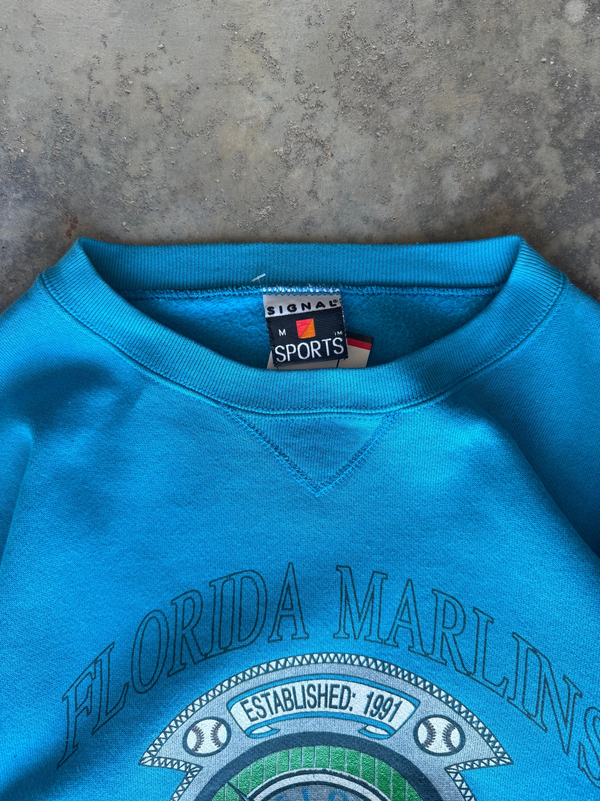 A close-up of a bright turquoise crewneck sweatshirt featuring a Florida Marlins graphic on the front and a visible Lionel Sports neck label, laid flat on a textured gray concrete surface.