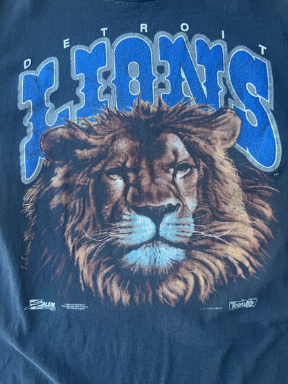 A close-up of a vintage Detroit Lions graphic t-shirt featuring a detailed lion face and the team name in blue and silver lettering on a navy blue fabric background.