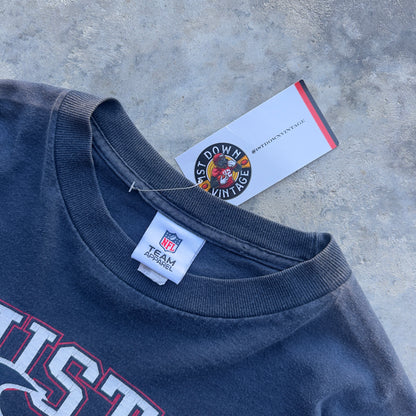 A close-up of a dark navy blue vintage football t-shirt with a crew neck, featuring a white NFL Team Official tag and a 'First Down Vintage' brand hangtag on a textured gray concrete surface.