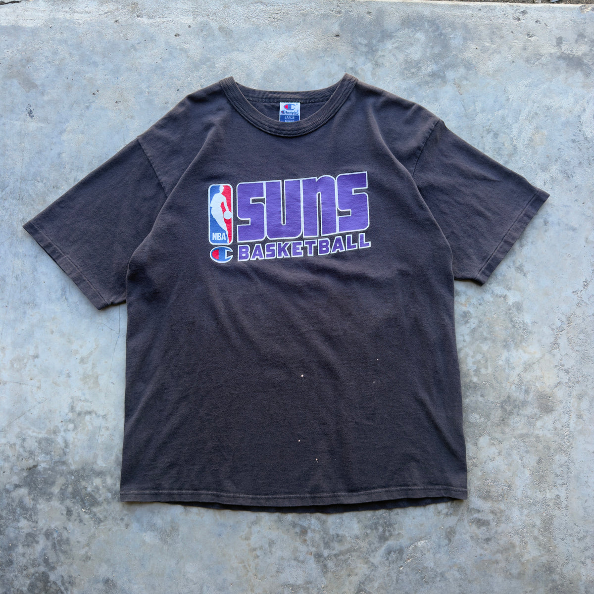 A faded black short-sleeve t-shirt featuring a purple and white Phoenix Suns Basketball graphic on the front, laid flat on a textured gray concrete surface.
