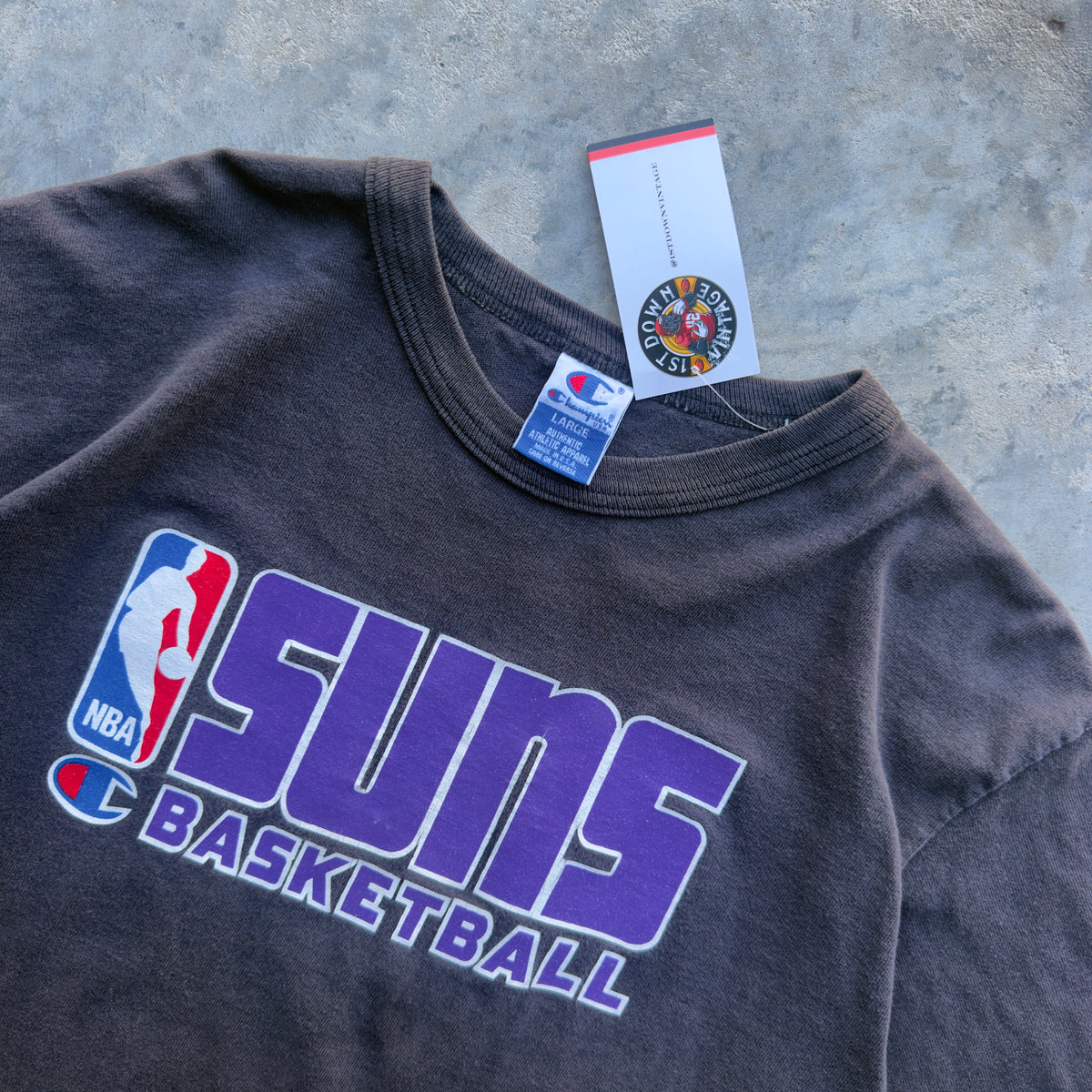 A dark charcoal gray Phoenix Suns basketball t-shirt with a purple, white, and blue graphic print on the front. The shirt is laid flat on a textured gray concrete surface, with its original retail tags visible at the collar.