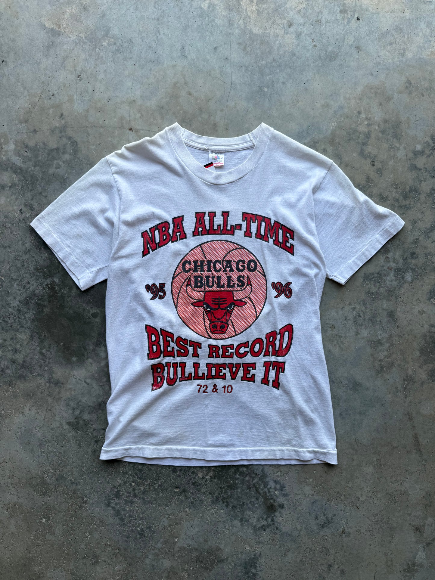 A white short-sleeve graphic T-shirt laid flat on a textured gray concrete surface. The front features a red and pink Chicago Bulls design with the text 'NBA ALL-TIME', 'CHICAGO BULLS', 'BEST RECORD BULLIEVE IT', and '72 & 10'.