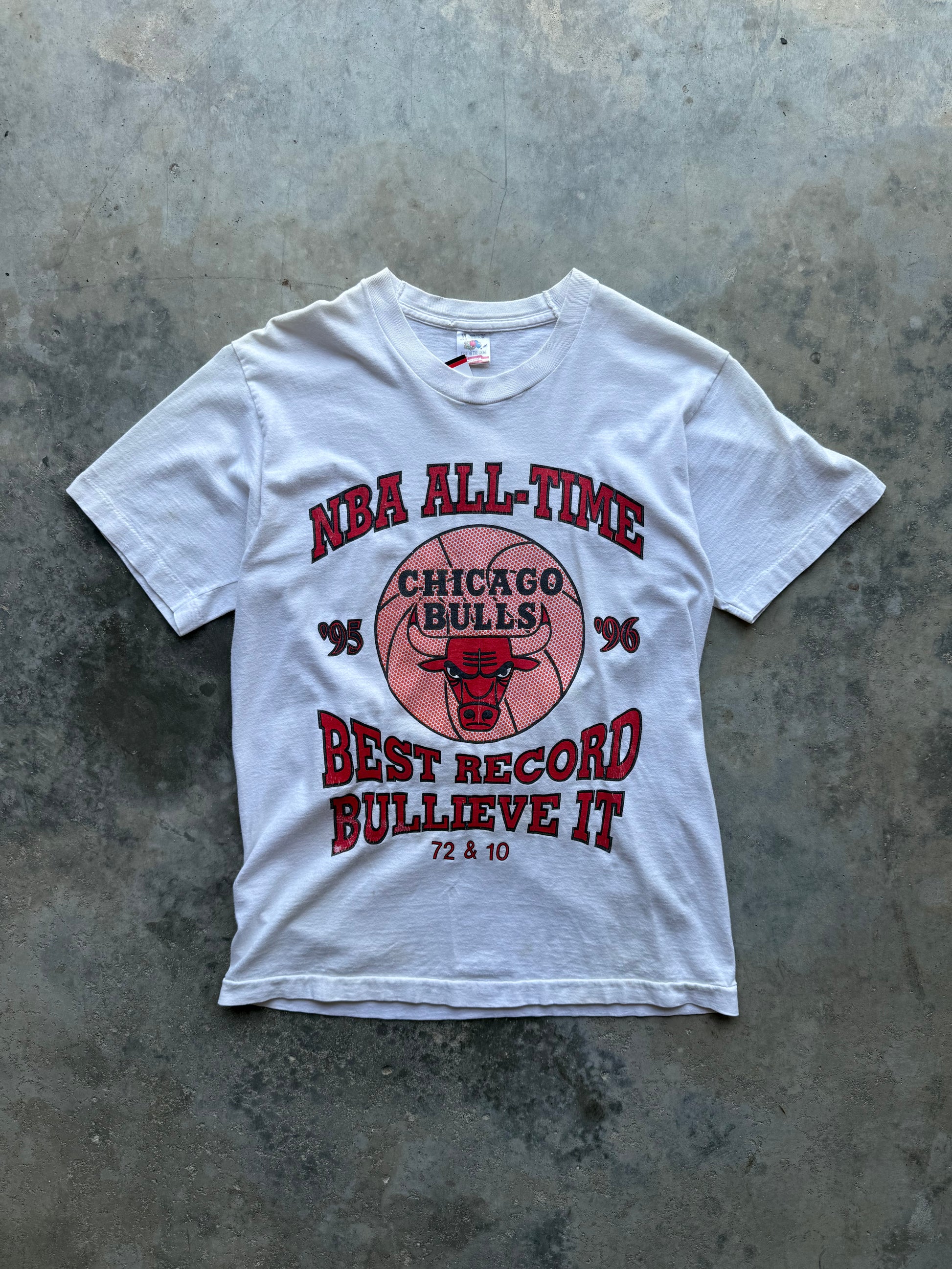 A white short-sleeve graphic T-shirt laid flat on a textured gray concrete surface. The front features a red and pink Chicago Bulls design with the text 'NBA ALL-TIME', 'CHICAGO BULLS', 'BEST RECORD BULLIEVE IT', and '72 & 10'.