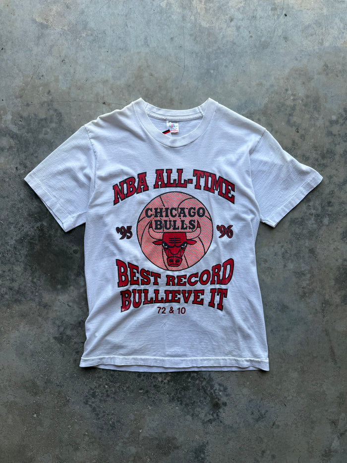 A white short-sleeve graphic T-shirt laid flat on a textured gray concrete surface. The front features a red and pink Chicago Bulls design with the text 'NBA ALL-TIME', 'CHICAGO BULLS', 'BEST RECORD BULLIEVE IT', and '72 & 10'.