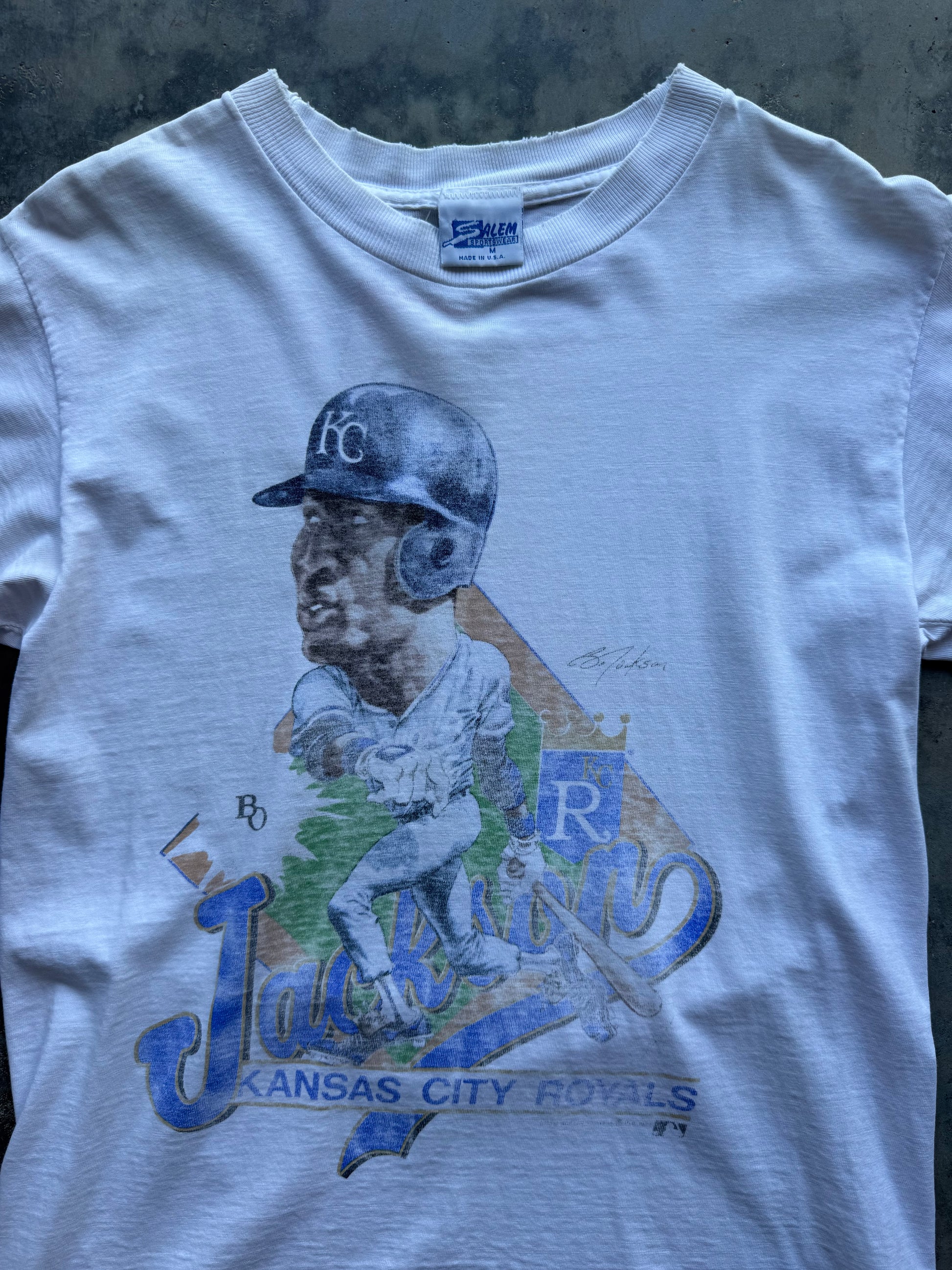 A white short-sleeve graphic t-shirt featuring a stylized illustration of baseball player Jackie Robinson with a blue helmet and 'Kansas City Royals' text, laid flat on a dark textured surface.