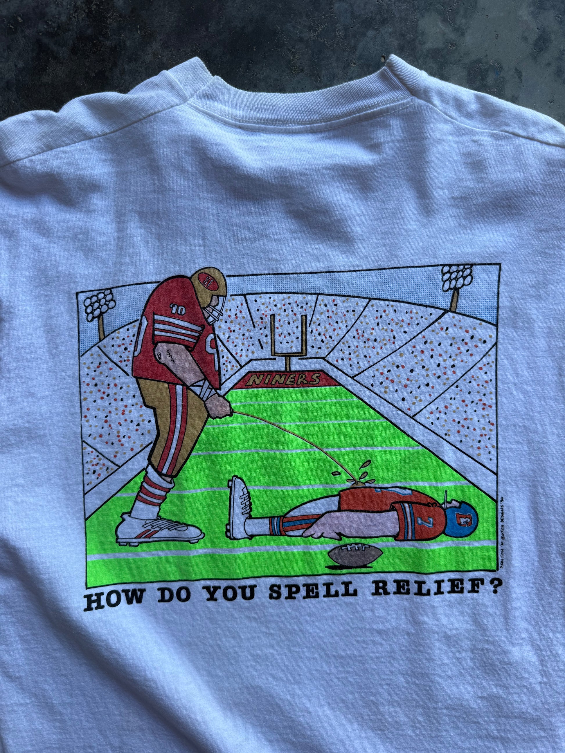 A light blue graphic t-shirt laid flat on a dark textured surface. The back of the shirt features a cartoon illustration of a San Francisco 49ers football player relieving another player on the field, with the text 'HOW DO YOU SPELL RELIEF?' printed below.